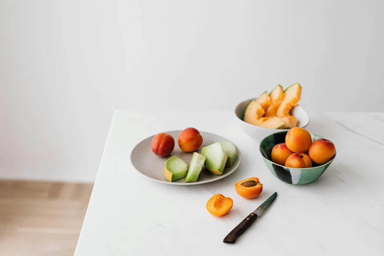 a table topped with bowls of fruit and a knife, by Nicolette Macnamara, unsplash, minimalism, matte white background, peach, organic ceramic white, background image