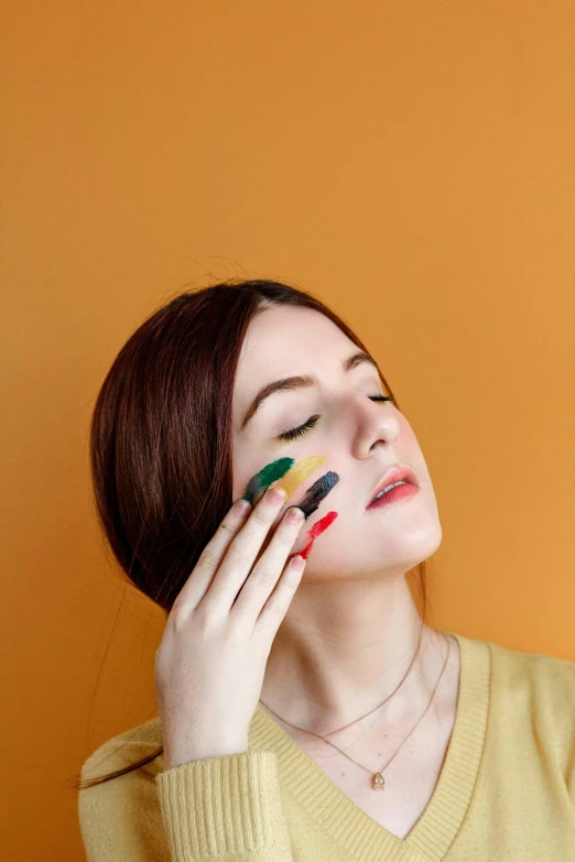 a woman with colorful paint on her face, an album cover, inspired by Elsa Bleda, trending on pexels, color field, hand on cheek, yellow and ornage color scheme, woman very tired, matte paint colors
