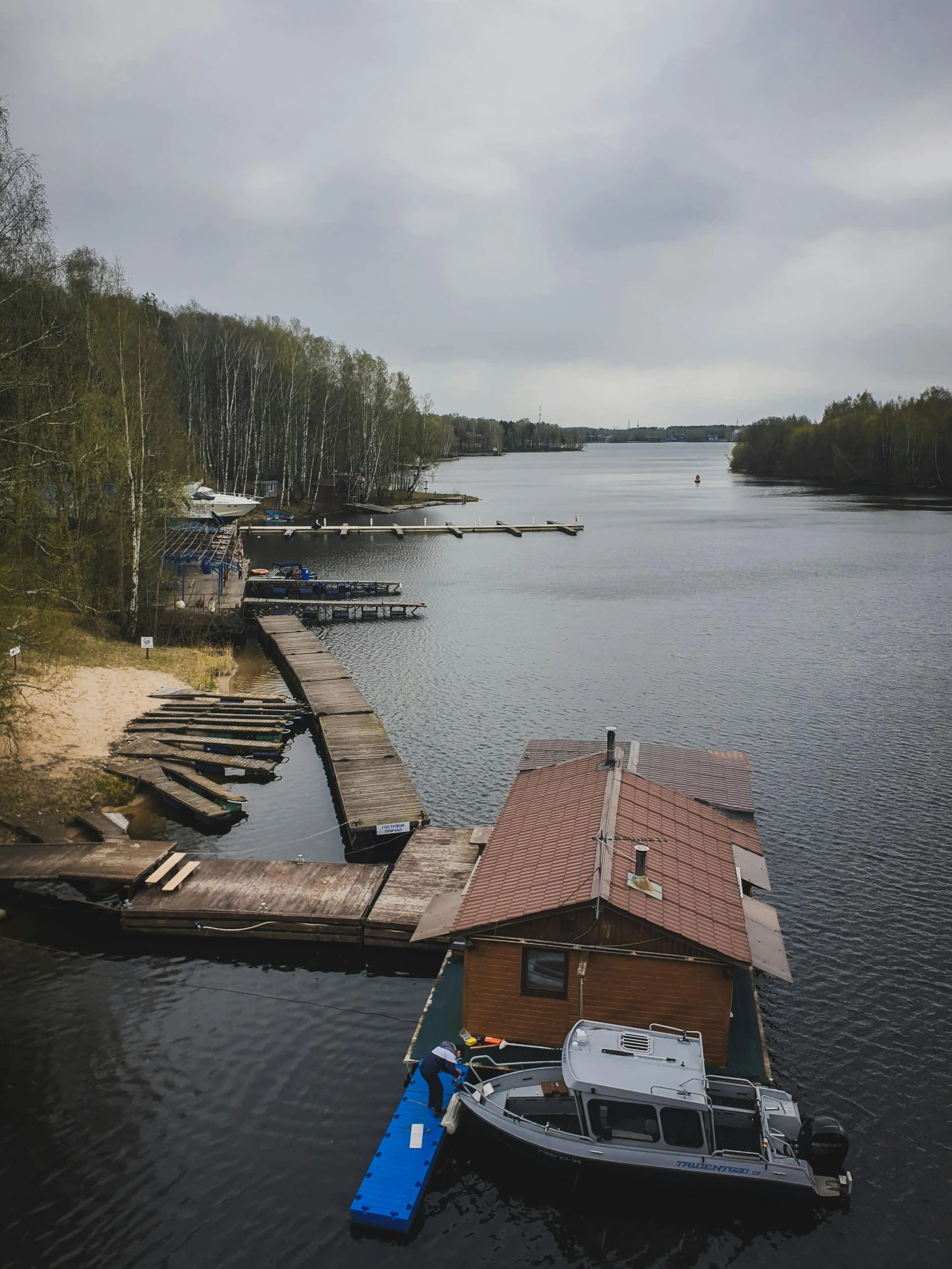 a boat sitting on top of a lake next to a dock, by Grytė Pintukaitė, hurufiyya, view from high, camps in the background, slight overcast lighting, looking towards the camera