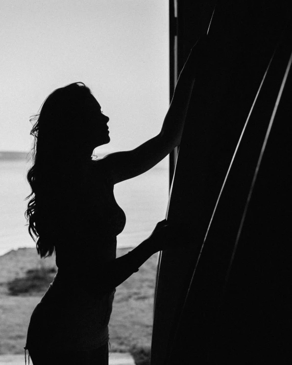 a woman standing in front of a window next to the ocean, a black and white photo, inspired by Max Dupain, unsplash, beautiful female body silhouette, leaning on door, anna nikonova aka newmilky, during a sunset