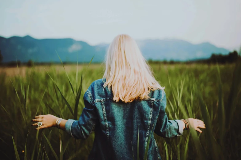 a woman standing in a field of tall grass, a picture, trending on unsplash, clean blonde hair, wearing a jeans jackets, hands behind back, mountains in the distance