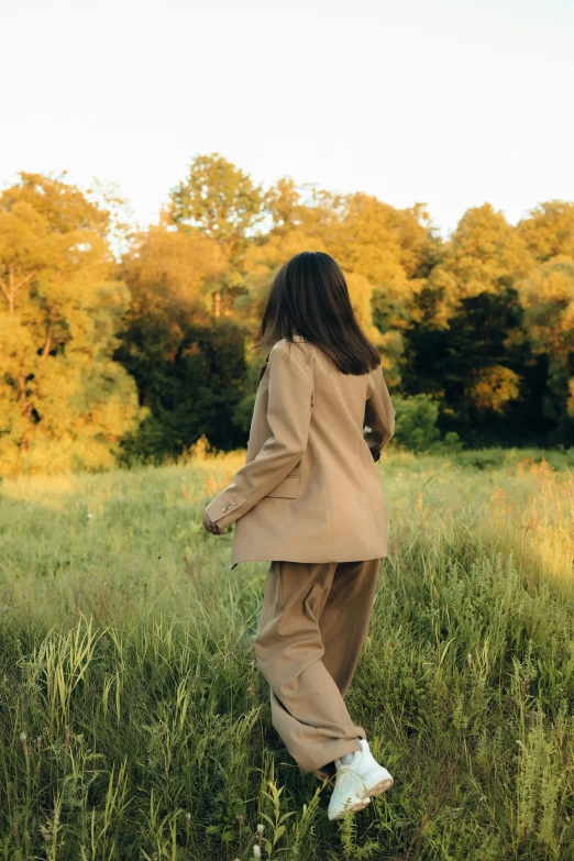 a woman walking through a field of tall grass, trending on pexels, tan suit, in a park, light brown coat, forest outside