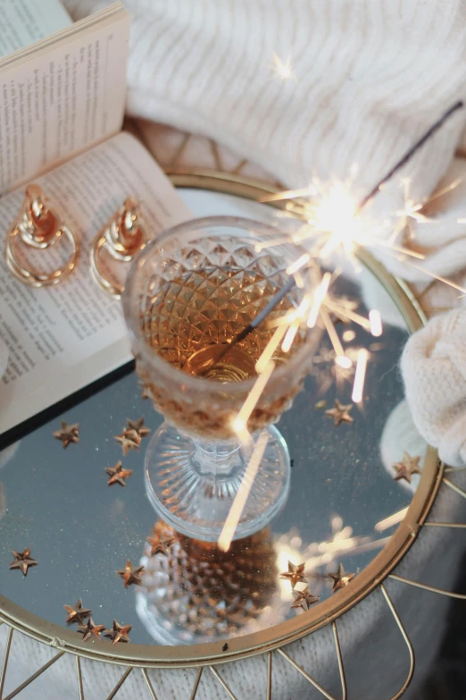 a person holding a sparkler on top of a table, a still life, by Julia Pishtar, trending on unsplash, glass with rum, glittering multiversal ornaments, reading for a party, glass - reflecting - stars