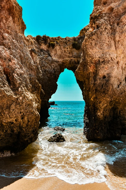 a couple of rocks sitting on top of a sandy beach, an archway, rocha, clear blue water, deep chasm