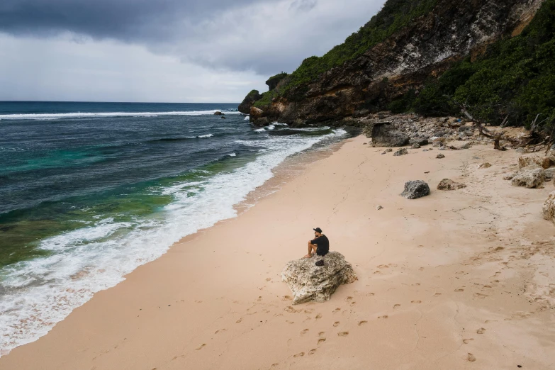 a person sitting on a rock on a beach, by Simon Marmion, pexels contest winner, at a tropical beach, lachlan bailey, slight overcast, conde nast traveler photo