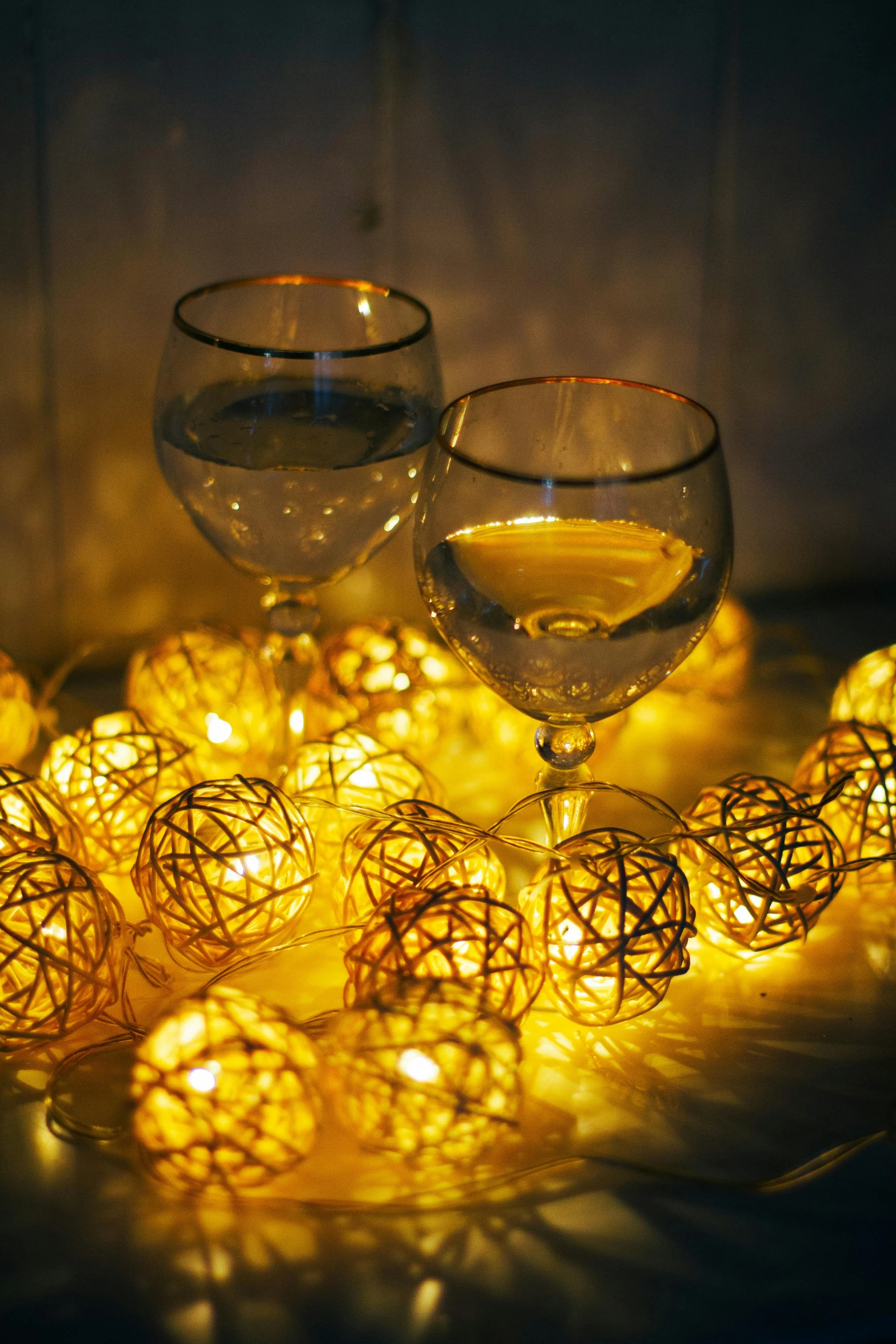 a couple of wine glasses sitting on top of a table, a still life, inspired by Patrick Dougherty, pexels, glowing lanterns, white lights, water reflecting suns light, gold decorations