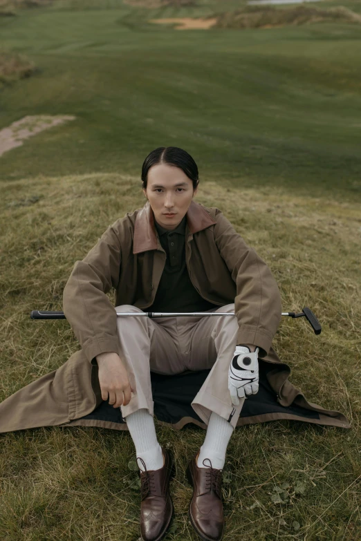 a woman sitting on top of a grass covered field, an album cover, inspired by Fei Danxu, he is wearing a trenchcoat, golf course, non binary model, looking straight to camera