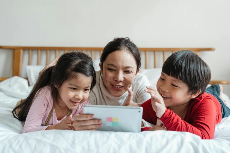 a woman and two children laying on a bed looking at a tablet, a cartoon, pexels contest winner, asian features, children's animated films, multicoloured, bedhead