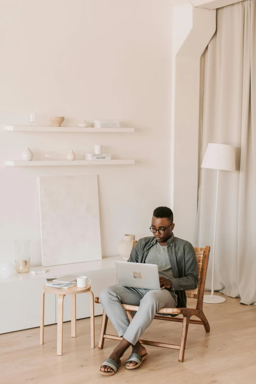 a man sitting in a chair using a laptop, a minimalist painting, trending on pexels, white room, black man, wearing a linen shirt, performing