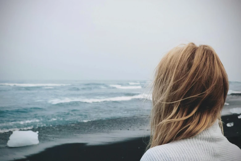 a woman standing on top of a beach next to the ocean, a picture, by Lucia Peka, trending on unsplash, romanticism, messy hair bedhead, she's sad, close up of a blonde woman, black sand