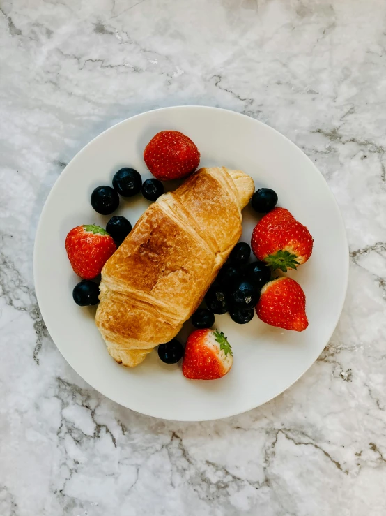 a croissant with strawberries and blueberries on a plate, by Kristin Nelson, unsplash, medium long shot, taken on iphone 14 pro, sleek white, wide high angle view