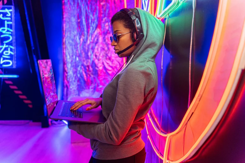 a woman wearing headphones using a laptop, interactive art, neon jungle, gaming room, girl wearing hoodie, multicoloured