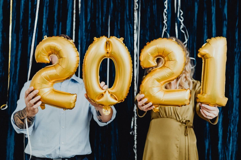 a man and a woman holding balloons in the shape of the numbers 2012, trending on pexels, happening, gold linens, background image, college party, puff sleeves