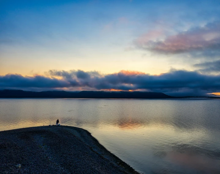 a person standing on a beach next to a body of water, by Jesper Knudsen, pexels contest winner, sunset panorama, mongolia, 2 people, grey