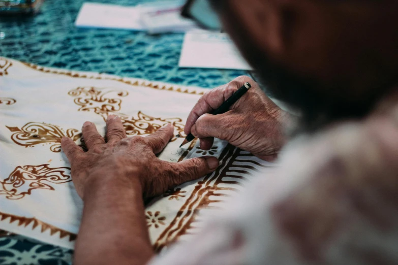 a person sitting at a table writing on a piece of paper, a silk screen, by Meredith Dillman, pexels contest winner, middle eastern style vendors, batik, afternoon time, gold embroidery