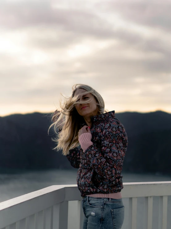 a woman standing on a balcony next to a body of water, a picture, inspired by Louisa Matthíasdóttir, trending on unsplash, wearing a fancy jacket, windy mood, standing on mountain, patterned clothing