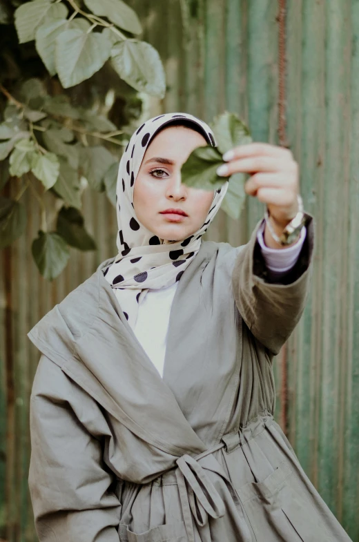 a woman holding a leaf in front of her face, by Ismail Acar, trending on pexels, hurufiyya, wearing a simple robe, prints, 2030s, grey