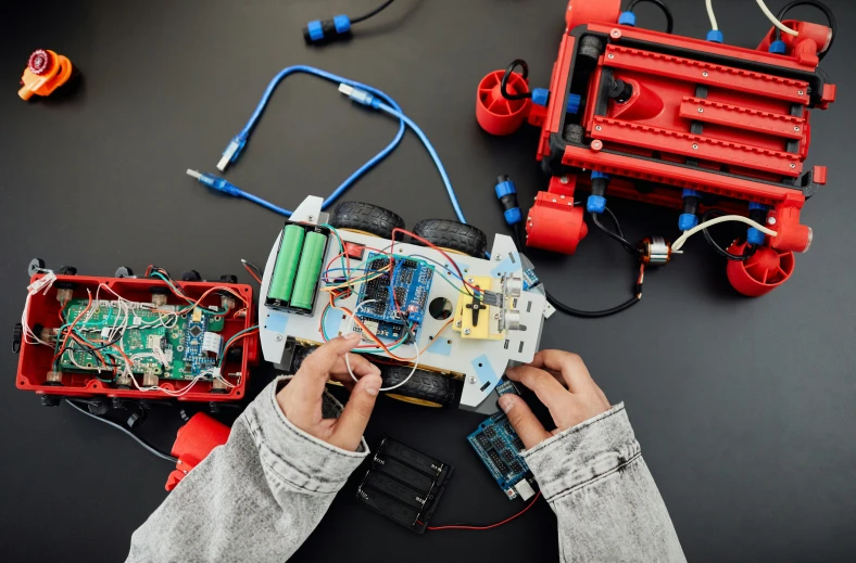 a person holding an electronic device on top of a table, pexels contest winner, process art, red and black robotic parts, red and cyan theme, only one robot kid on the ground, circuits