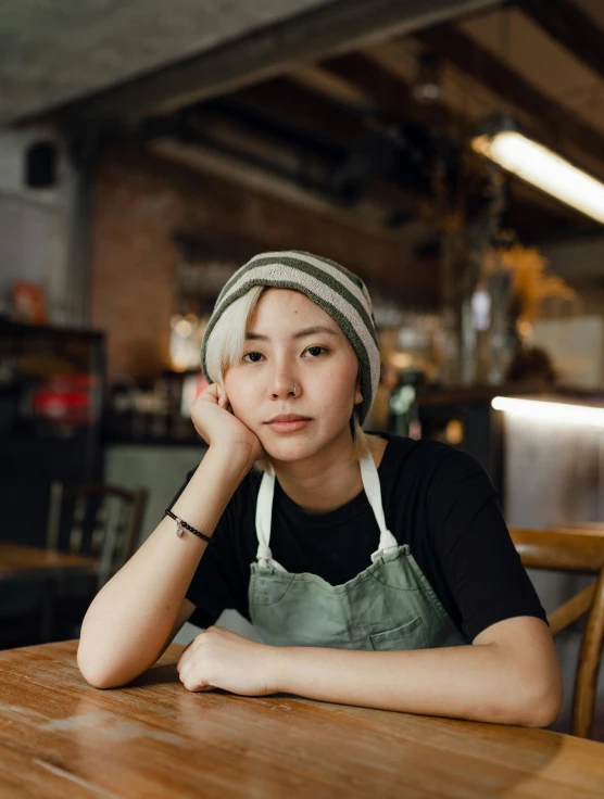 a woman sitting at a table in a restaurant, a portrait, inspired by Yukimasa Ida, trending on unsplash, wearing beanie, wearing an apron, non binary model, portrait”