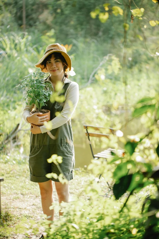 a woman in a hat holding a potted plant, a green gold forest in japan, wearing overalls, ecovillage, soft shade