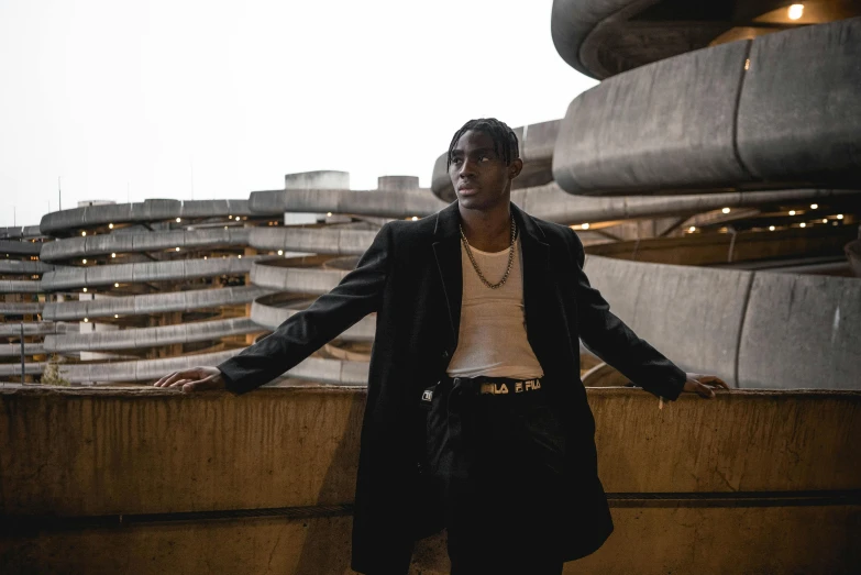 a man standing on a ledge in front of a building, an album cover, by Cosmo Alexander, pexels contest winner, he is wearing a black trenchcoat, lil uzi vert, elegant cinematic pose, standing in an arena