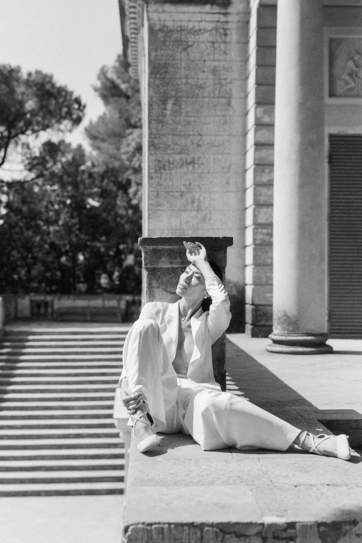 a man sitting on the steps of a building, a black and white photo, bauhaus, wearing white pajamas, blocking the sun, roman shipunov, woman posing