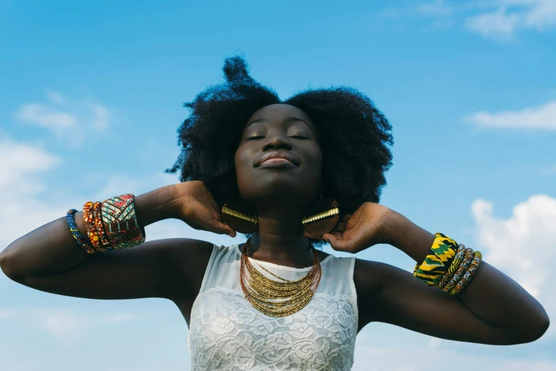 a woman standing in a field with her hands on her head, inspired by Ras Akyem, pexels contest winner, afrofuturism, bracelets and necklaces, blue sky, ( ( dark skin ) ), afro