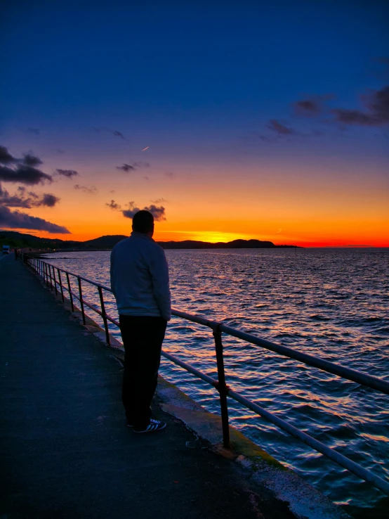 a man standing on top of a pier next to the ocean, by Alison Watt, pexels contest winner, sunsetting color, profile image, holywood scene, looking off to the side