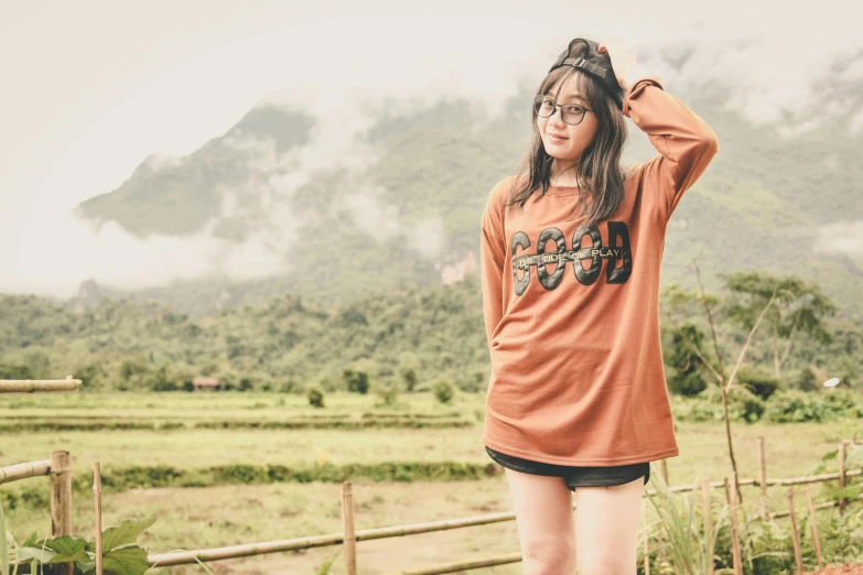 a woman standing on top of a lush green field, inspired by Ruth Jên, pexels contest winner, long orange sweatshirt, girl wearing round glasses, caramel. rugged, graphic tees