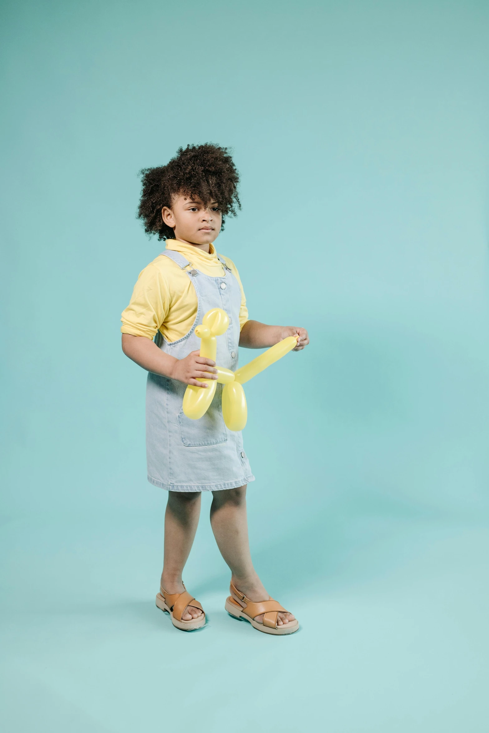 a little girl standing in front of a blue background, inspired by Sarah Lucas, shutterstock contest winner, yellow overall, duck shoes, happy fashion model, in pastel shades