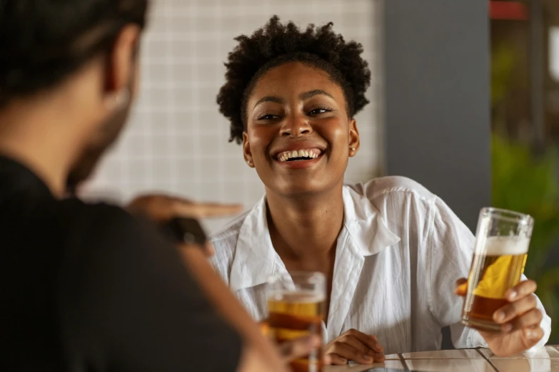 a woman sitting at a table with a glass of beer, brown skin man with a giant grin, wearing a white button up shirt, drinking whiskey, flirting smiling