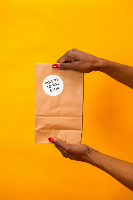 a person holding a brown paper bag with a sticker on it, inspired by Géza Dósa, back to camera, dark skinned, serving suggestion, on a yellow canva