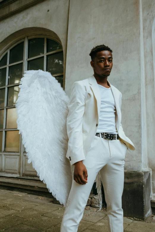 a man in a white suit standing in front of a building, an album cover, inspired by Louis Hersent, pexels contest winner, renaissance, big angel wings on back, black teenage boy, model shoot, cupid