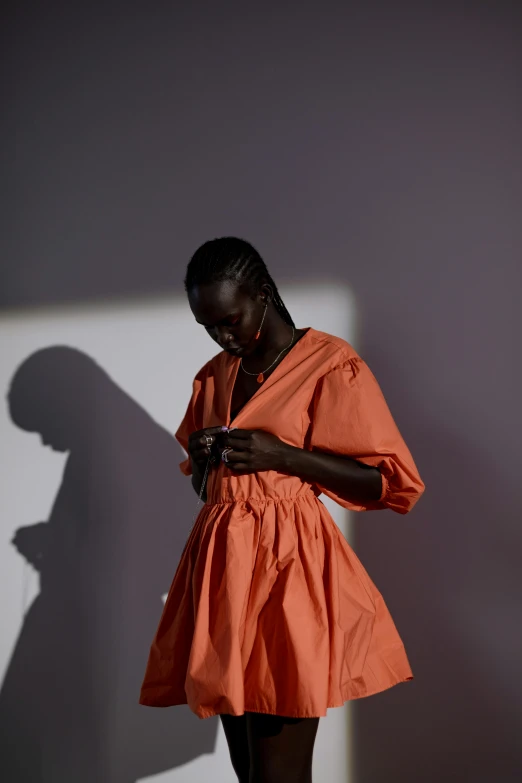 a woman in an orange dress standing against a wall, inspired by Georges de La Tour, trending on pexels, adut akech, mid morning lighting, issey miyake, grey orange