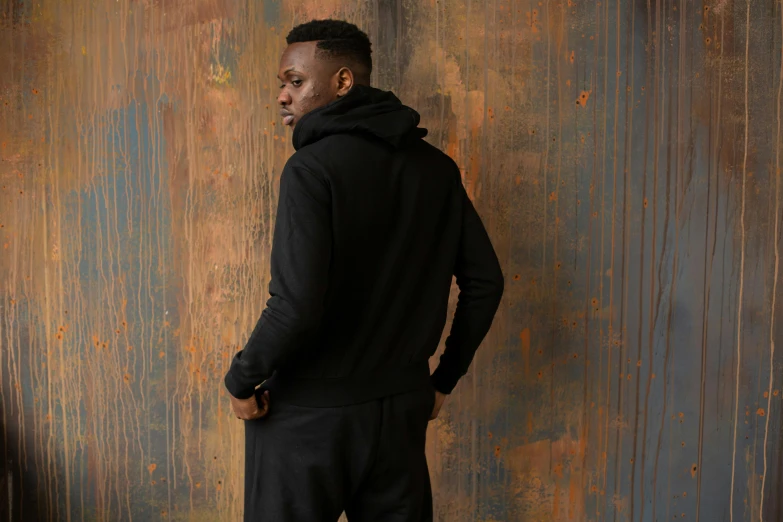 a man standing in front of a rusty wall, inspired by Paul Georges, dressed black hoodie, wearing a tracksuit, profile image, back view. nuri iyem