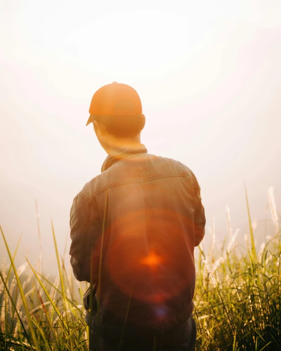 a man standing on top of a lush green field, trending on unsplash, sumatraism, fading rainbow light, wearing sunglasses and a cap, ethereal back light, sun behind her