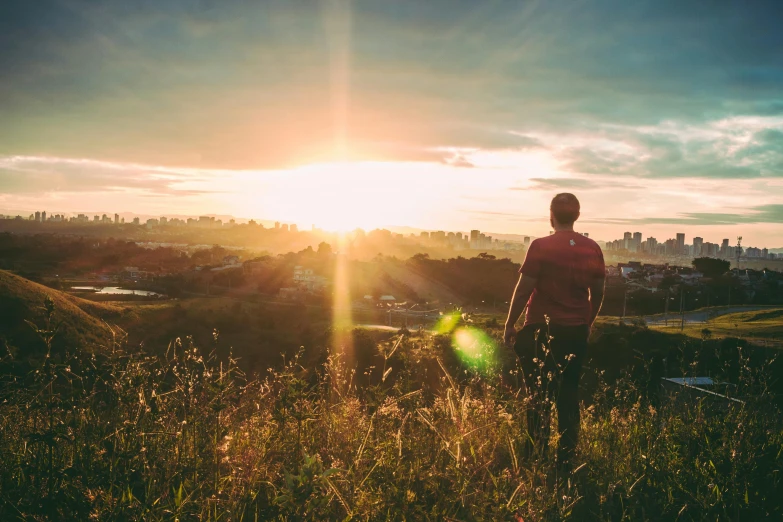 a man standing on top of a lush green hillside, pexels contest winner, sun behind her, sydney park, grass field surrounding the city, a photo of a man