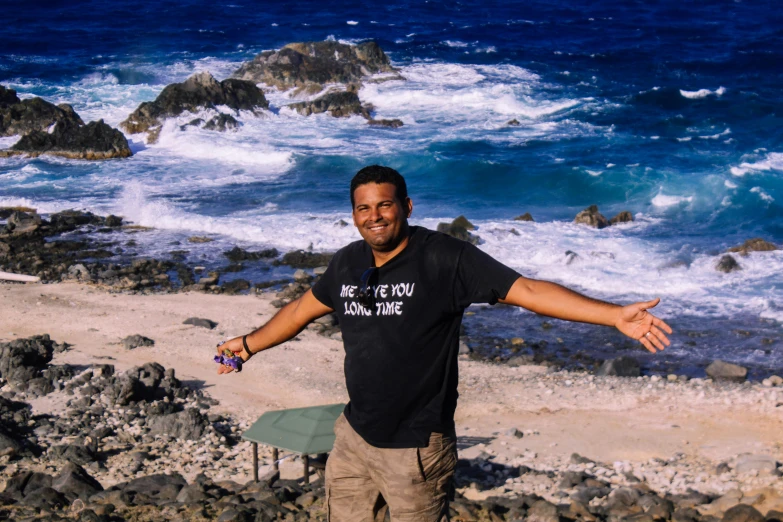 a man standing on a rocky beach next to the ocean, happening, avatar image, mutahar laughing, samoan features, watch photo