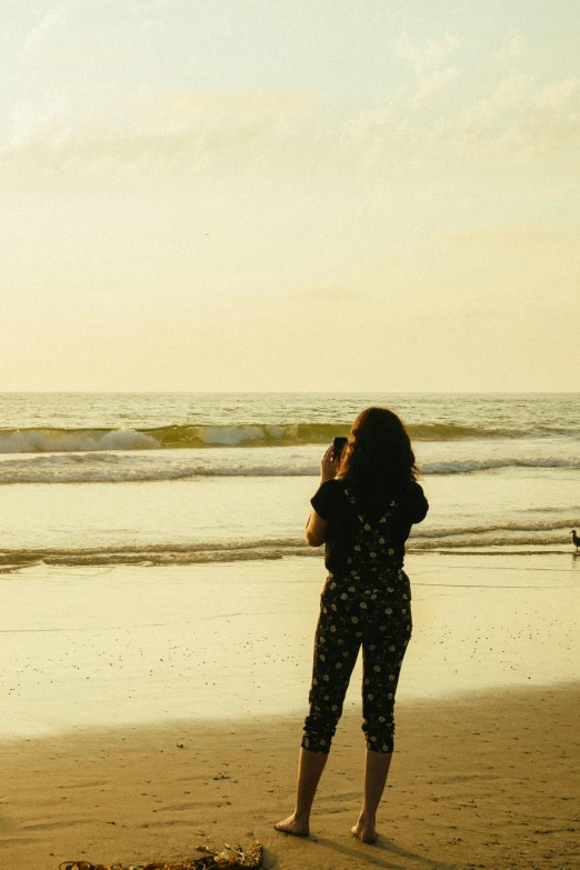 a woman standing on top of a beach next to the ocean, taking a picture, late afternoon, facing away, beaches