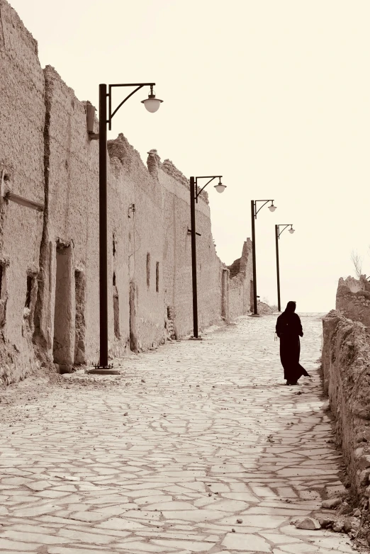 a black and white photo of a person walking down a cobblestone street, inspired by Osman Hamdi Bey, white desert, city wall, sepia colors, alone!!