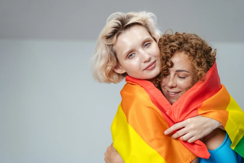a woman holding a child wrapped in a rainbow blanket, a photo, shutterstock, antipodeans, short curly blonde haired girl, lgbt flag, on grey background, her friend