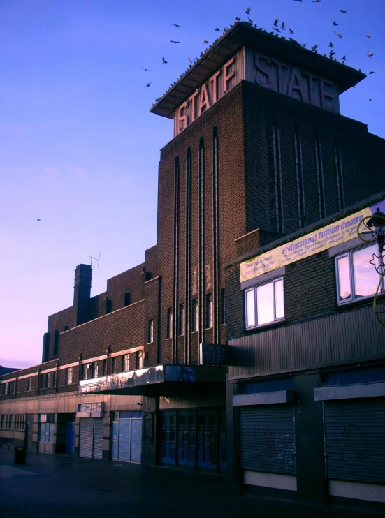 a building with a lot of birds flying around it, a colorized photo, by Steve Prescott, unsplash, brutalism, an old cinema, elstree, front view 1 9 9 0, sun down