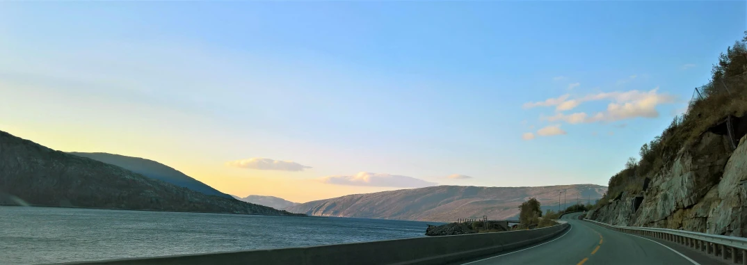 a car driving down a road next to a body of water, a picture, over the hills, blue sky at sunset, yggrdasil, fjord