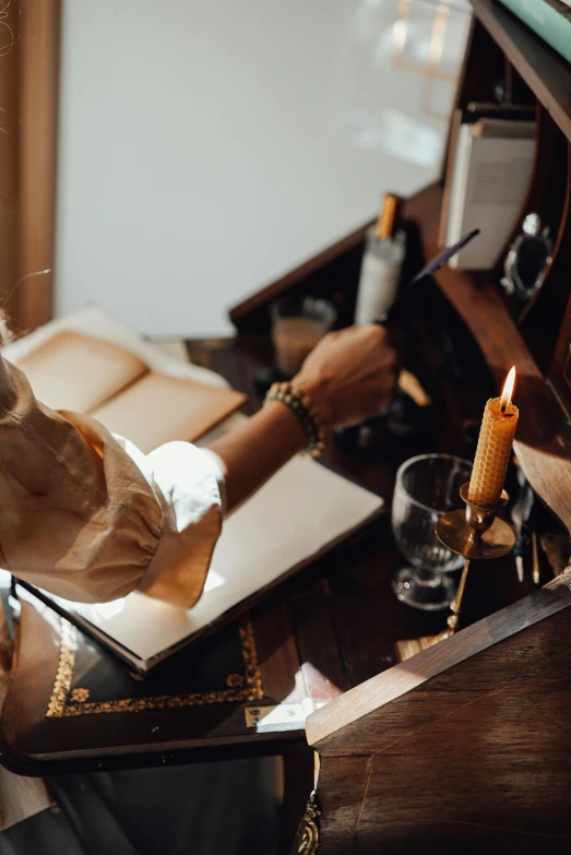 a woman sitting at a desk writing on a piece of paper, by Julia Pishtar, waxy candles, leather cuffs around wrists, champagne on the table, crafting spells