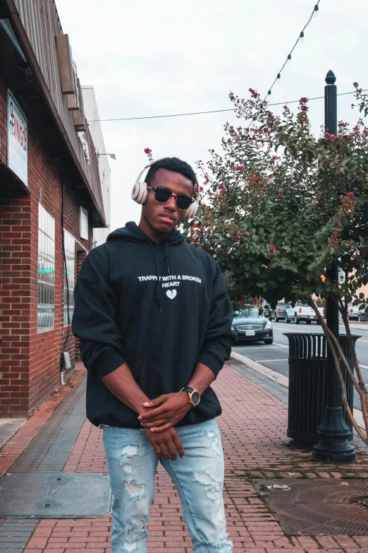 a man standing on a sidewalk in front of a building, an album cover, by Everett Warner, trending on unsplash, hoodie, he is from virginia, wearing black headphones, posing for a picture