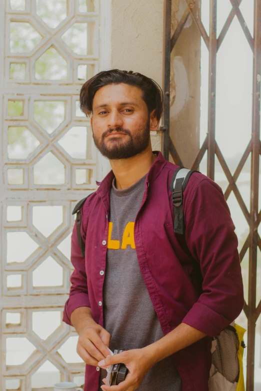 a man standing next to a wall holding a camera, inspired by Jitish Kallat, smirking at the camera, profile image, wearing casual clothing, islamic
