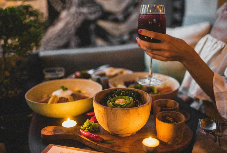 a woman sitting at a table with a glass of wine, pexels contest winner, bowl filled with food, on a wooden tray, holding a candle holder, cheers