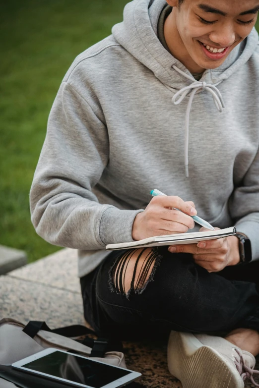 a man sitting on the ground writing on a notebook, inspired by John Luke, trending on unsplash, academic art, gray hoodie, teenage girl, non-binary, professional grade