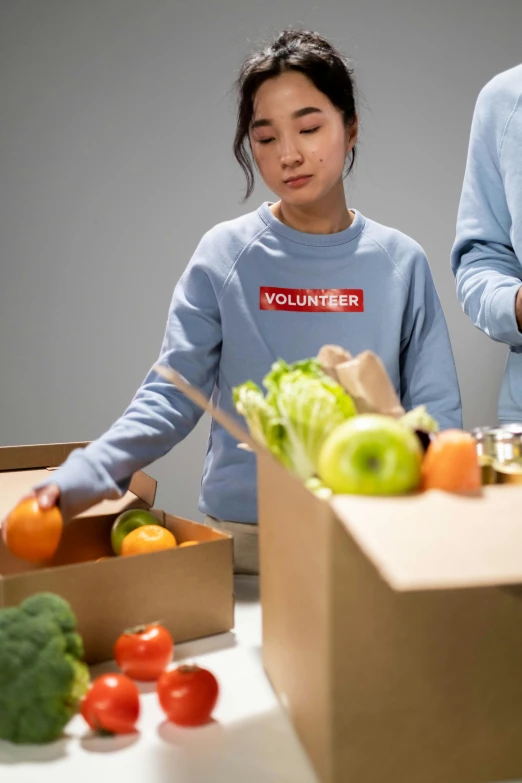 a couple of people standing next to a box of food, a picture, on a gray background, sustainability, a young asian woman, lights on