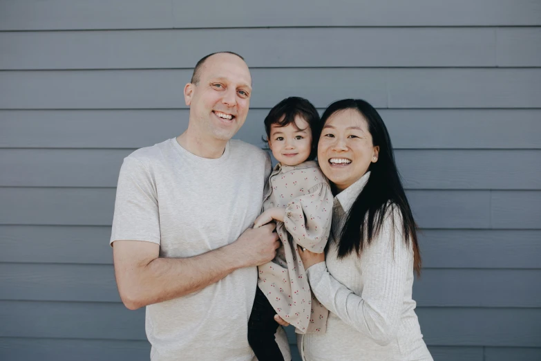 a man and woman holding a child in front of a gray wall, a picture, unsplash, mingei, tyler edlin and natasha tan, in front of white back drop, square, standing outside a house